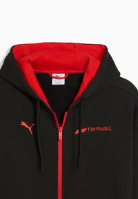 Crni dukserica na zatvaranje s crvenom unutrašnjošću, s crvenim Puma logotipom i natpisom "Formula 1". Uže za zatezanje i patent zatvarač u kontrastnim bojama.