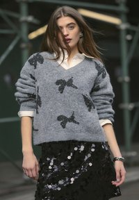 Pullover grigio a maglia con fiocchi di paillettes nere, abbinato a una gonna nera con paillettes. L'outfit è completato da una camicia bianca con colletto a strati sotto.