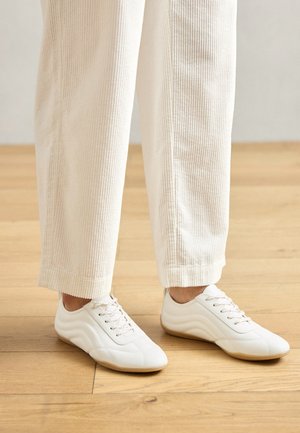 Baskets blanches avec une texture en cuir lisse, un système de laçage et un design contour. Portées avec un pantalon en velours côtelé beige clair.