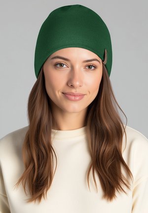 Bonnet en tricot vert à texture lisse avec une étiquette en cuir sur le côté, porté avec un sweat-shirt couleur crème. Gros plan sur le visage et les cheveux.