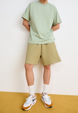 Personne portant un t-shirt Nike vert clair, un short beige, des chaussettes blanches et des baskets Nike blanches avec des accents orange, se tenant sur un sol jaune.