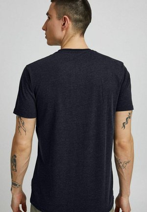 Mann mit kurzen dunklen Haaren, von hinten gesehen, trägt ein einfaches dunkelgraues T-Shirt, zeigt Tattoos auf beiden Unterarmen.