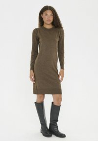 Braunes Strickkleid mit langen Ärmeln und gerippten Bündchen, ergänzt durch schwarze kniehohe Stiefel. Weiche Textur und figurbetonter Schnitt.