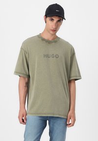 Olívovo zelené oversized tričko s nápisom "HUGO" v sivej farbe, s okrúhlym výstrihom a viditeľnými švami, skombinované s svetlo modrými džínsami.