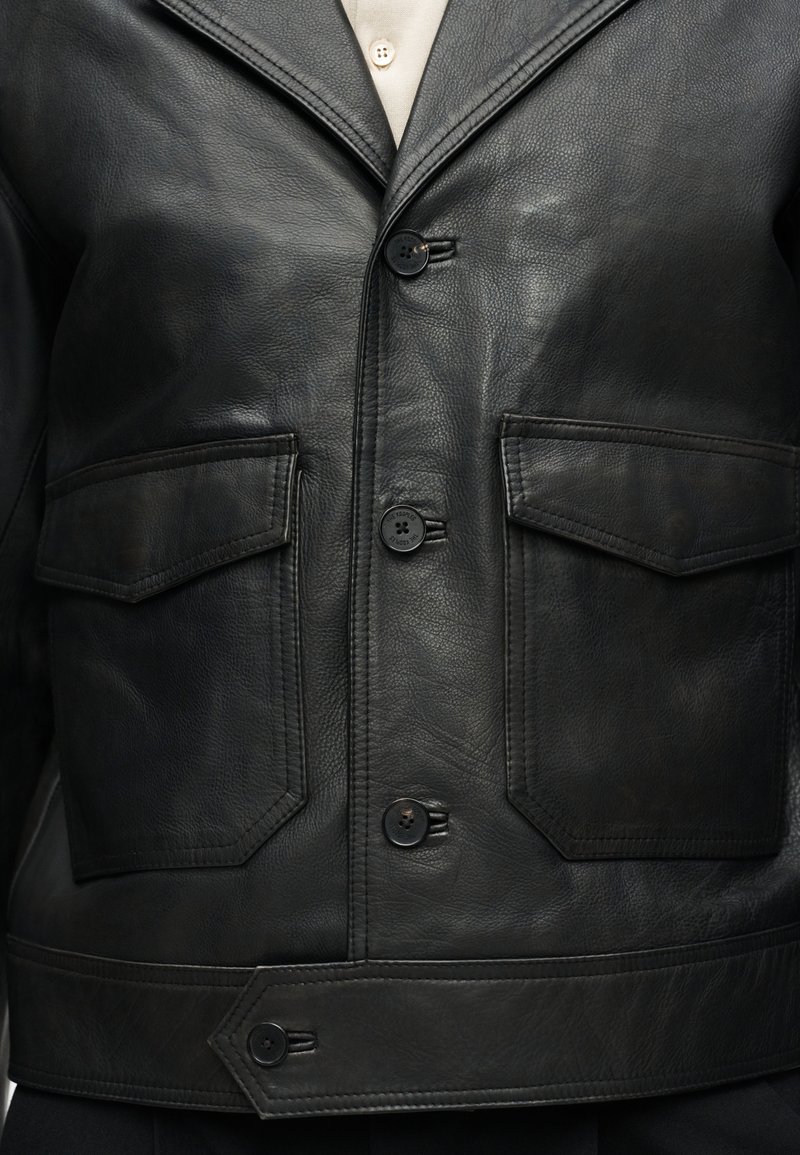 Black leather jacket with front buttons, two flap pockets, and visible white shirt collar underneath.