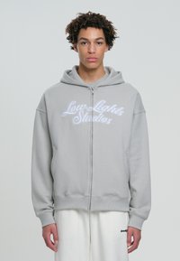 Felpa grigia con zip, con un grande logo ricamato "Low Lights Studios" in bianco, dotata di tasca frontale e una morbida texture di tessuto.