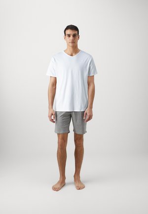 Hombre joven de pie descalzo con una camiseta blanca lisa de manga corta y pantalones cortos grises hasta la rodilla, frente a un fondo sencillo.