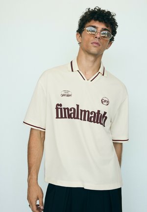Junger Mann mit lockigem Haar, der ein beiges Polohemd mit dem Text "final match" und Sonnenbrille trägt, steht vor einem schlichten weißen Hintergrund.