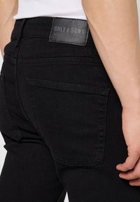 Vaqueros de mezclilla negra con un ajuste slim, costuras sutiles en los bolsillos y una etiqueta de cuero texturizado en la parte posterior, con la marca "ONLY & SONS."