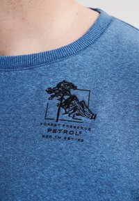 Sudadera de algodón azul con cuello de canalé, que presenta un gráfico bordado en negro de un árbol y montañas, junto con el texto "RESERVA DEL BOSQUE".