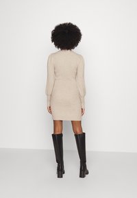 Stickad beige klänning med långa ärmar och ribbad textur, med en figurnära silhuett och rynkade axeldetaljer, kombinerad med höga svarta stövlar.