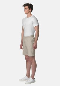 T-shirt bianco, pantaloni beige con tasche laterali e sneakers bianche. Il tessuto appare liscio; i pantaloni hanno una vita morbida e un sottile dettaglio con logo.