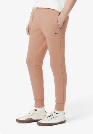 Personne portant un pantalon de jogging slim beige avec des poches et des baskets blanches avec des semelles marron, debout sur un fond blanc.