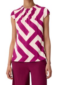 Blusa viola e crema con un motivo geometrico a zigzag, maniche corte a cappuccio e un dettaglio di scollatura arricciata. Tessuto morbido e leggero.