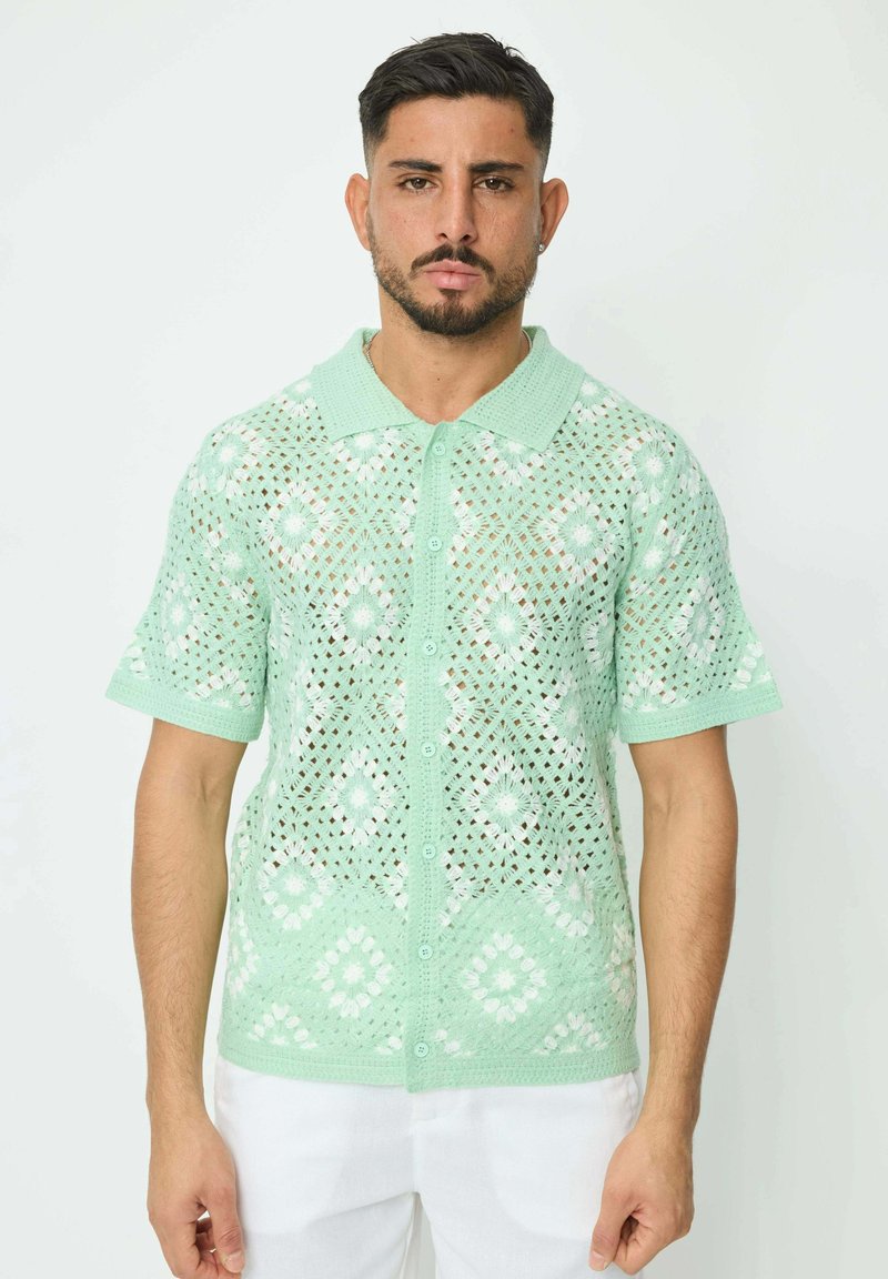 Polo en maille vert menthe, à manches courtes, avec un motif en losanges, des accents floraux blancs et une fermeture à boutons.