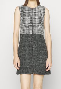 Robe sans manches avec un corsage à motifs en damier noir et blanc, une jupe en tissu noir et une fermeture éclair à l'avant, présentant une silhouette ajustée.