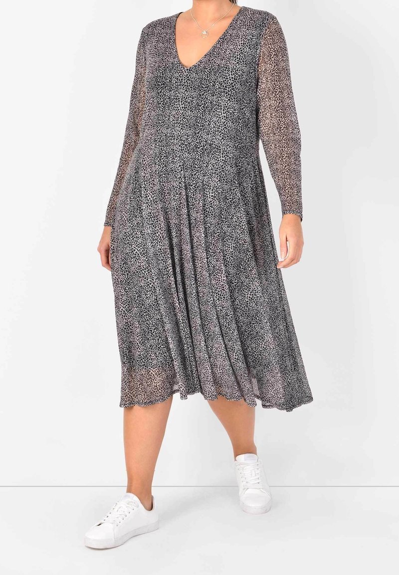 Langärmliges Kleid mit V-Ausschnitt, das mit einem schwarz-grauen Leopardenmuster, fließendem Stoff und einem plisseeartigen Rock gestaltet ist, kombiniert mit weißen Turnschuhen.