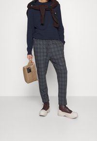 Marinblå tröja, grårutiga byxor och kraftiga bruna snörstövlar. En brun väska med en etikett hålls i vänster hand.