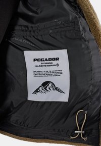Giacca in pile marrone con fodera nera e un tag etichettato. Il tag include grafiche montane e istruzioni per la cura.