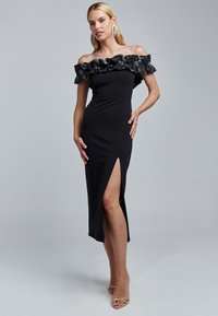 Black off-the-shoulder dress featuring a textured ruffled neckline, fitted bodice, and thigh-high slit, made from smooth fabric.