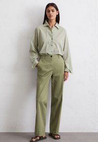 Light green oversized button-up shirt with a collar, paired with high-waisted light olive trousers and brown flat sandals. Smooth fabric and relaxed fit.