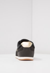 Svart träningsskor med en slät matt yta och en glänsande guldaccent vid hälen. Rundad form och kontrasterande vit sula.