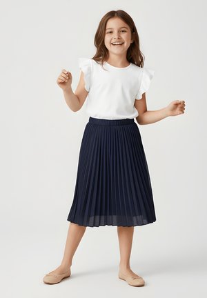 Uśmiechnięta dziewczyna ubrana w biały top z falbanami na rękawach, granatową plisowaną spódnicę midi i beżowe baleriny, stojąca z lekko uniesionymi ramionami.