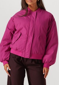 Fuchsia bomberjack met hoge kraag, elastische manchetten en taille. Voorzien van zijzakken en een gladde, licht gekreukte textuur.