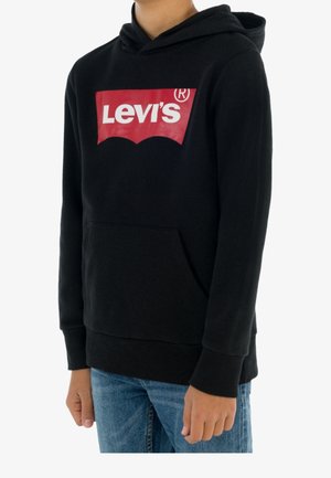 Person iført en sort Levi's hættetrøje med et rødt og hvidt logo, kombineret med blå jeans, stående foran en hvid baggrund.