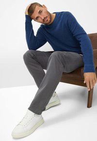 Blauwe gebreide trui, grijze broek en witte sneakers gedragen door een zittende man. Hij raakt zijn hoofd aan, met een bruine stoel zichtbaar op de achtergrond.