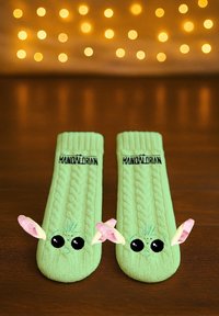 Green knit socks featuring cable design, large black eyes, and pink ears, with "MANDALORIAN" logo at the top.