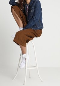 Giacca di jeans abbinata a culottes di velluto a coste marrone, sneakers bianche spesse e una maglia a righe. Modella seduta su uno sgabello bianco.