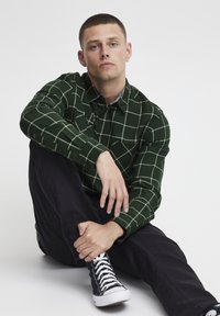 Cămașă cu carouri verzi și linii mai deschise de verde, cu două buzunare la piept, asortată cu pantaloni negri și adidași negri.