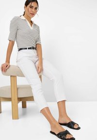 Gestreiftes Kurzarm-Polohemd in Weiß und Marineblau, kombiniert mit geraden weißen Hosen und schwarzen flachen Sandalen, sitzend auf einem beigen Hocker.