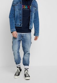 Hombre vestido con una chaqueta de mezclilla azul, sudadera navy con texto colorido, jeans de mezclilla claros, calcetines blancos y zapatillas altas negras.