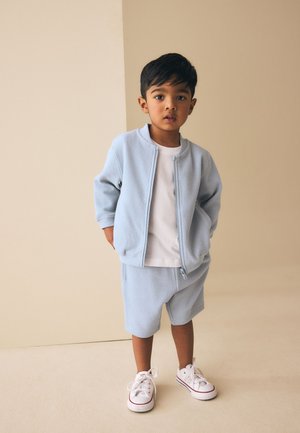 Ung dreng iført lyseblå, struktureret jakke og shorts med hvid t-shirt og hvide sneakers står foran en beige baggrund.