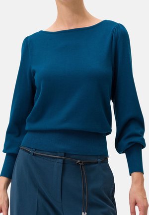 Vrouw draagt een turquoise blouse met lange mouwen en ballonmouwen, gecombineerd met bijpassende broek die is gestrikt met een dunne zwarte riem.