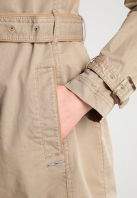 Beige trenchcoat met een riem, knoopmanchetten en een zijzak met een zilveren logo-tag. Gemaakt van soepel, duurzaam materiaal.