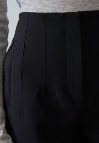 Pantaloni neri a vita alta con pieghe frontali, tasche laterali e un tessuto liscio e testurizzato. Abbinati a una maglia grigia.