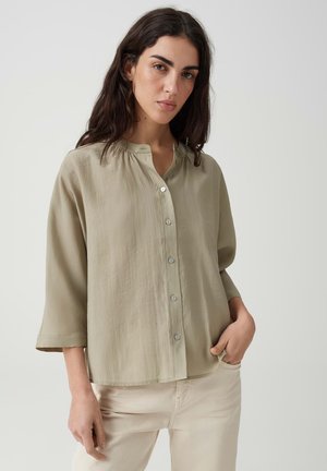 Femme aux longs cheveux foncés porte une chemise beige ample à boutons et un pantalon clair, debout devant un fond gris uni.