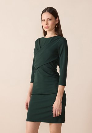Dunkelgrünes Kleid aus weichem Stoff, mit Wickeldesign, Dreiviertelärmeln und einer figurbetonten Silhouette mit gerafften Seitendetails.