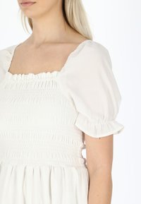 Witte jurk met vierkante halslijn en smock-bodice. Heeft pofmouwen met een gerimpelde rand en een lichte, luchtige textuur.