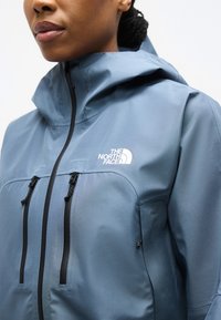 Femme portant une veste imperméable bleue avec capuche, comprenant deux fermetures éclair verticales sur la poitrine et le logo The North Face sur le côté gauche.