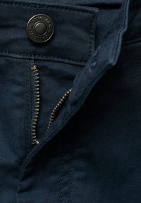 Temno modri džins hlače z metalnim zadrgo in okroglo črno gumbom na pasu. Tkanina prikazuje fino teksturo in podrobnosti šivanja.