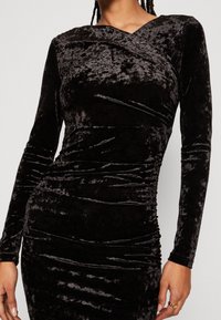 Robe en velours noir à manches longues, dotée d'un décolleté en V profond et de fronces le long du corps pour une silhouette ajustée. Texture lisse.