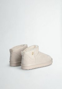 Stivaletti alla caviglia in suede beige con rivestimento soffice, bordo superiore curvo e dettaglio con logo sul lato. Presentano una suola in gomma testurizzata per una migliore aderenza.