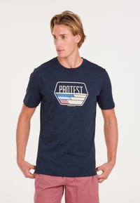 Camiseta de algodón azul marino con un logo hexagonal de "PROTEST" que presenta gráficos de olas coloridos en rosa y azul. Manga corta, cuello redondo.