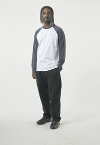 Langärmliges Raglan-T-Shirt mit weißem Körper und dunkelgrauen Ärmeln, kombiniert mit lockeren schwarzen Hosen und schwarzen Schuhen. Einfaches Logo auf der Brust sichtbar.