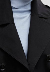 Manteau noir à double boutonnage porté par-dessus un pull à col roulé côtelé bleu clair, montrant la partie supérieure de la poitrine et du col.