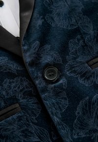 Blazer en velours bleu foncé avec un motif floral subtil, bouton noir, revers noir et une chemise blanche en dessous.
